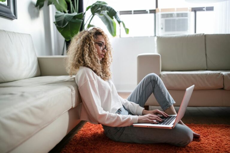 Woman sitting on the floor with a laptop in her lap typing with a blank expression on her face.