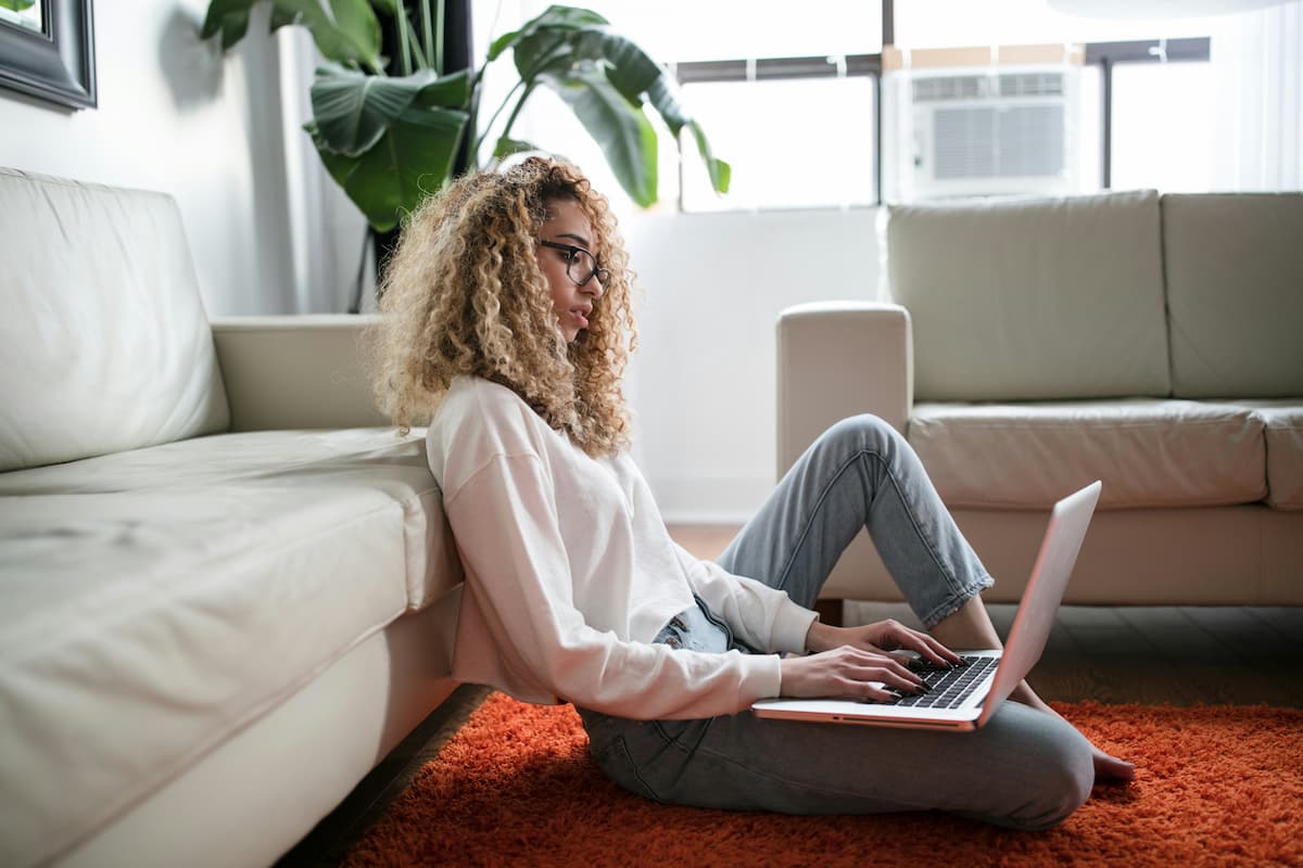 Woman sitting on the floor with a laptop in her lap typing with a blank expression on her face.